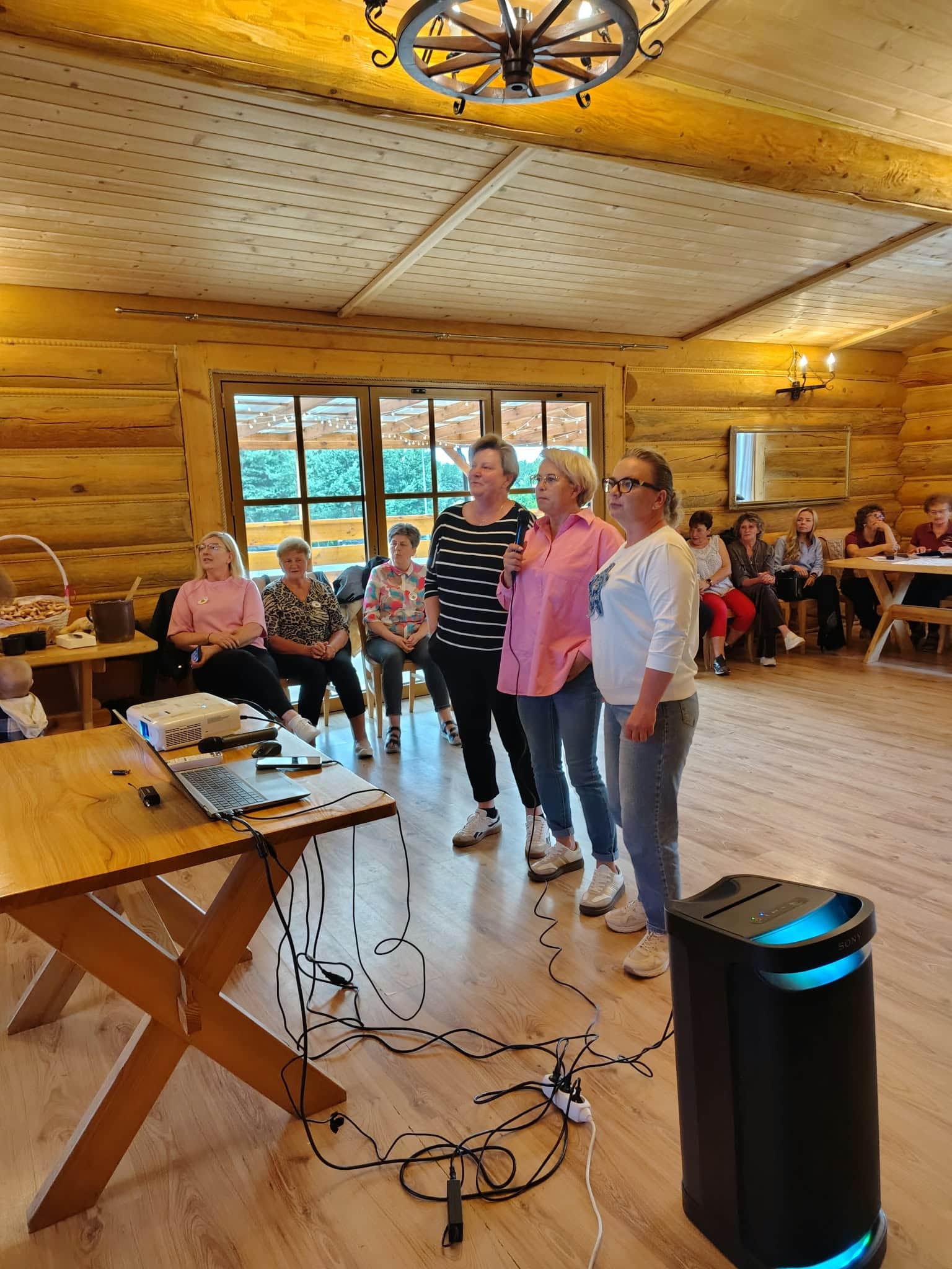 Grupa uczestniczek z mikrofonem przy sprzęcie do karaoke w drewnianej sali.